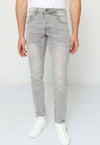 Jean en denim gris clair avec une coupe slim, comportant cinq poches et un léger effet décoloré. Associé à des baskets blanches sur un fond neutre.