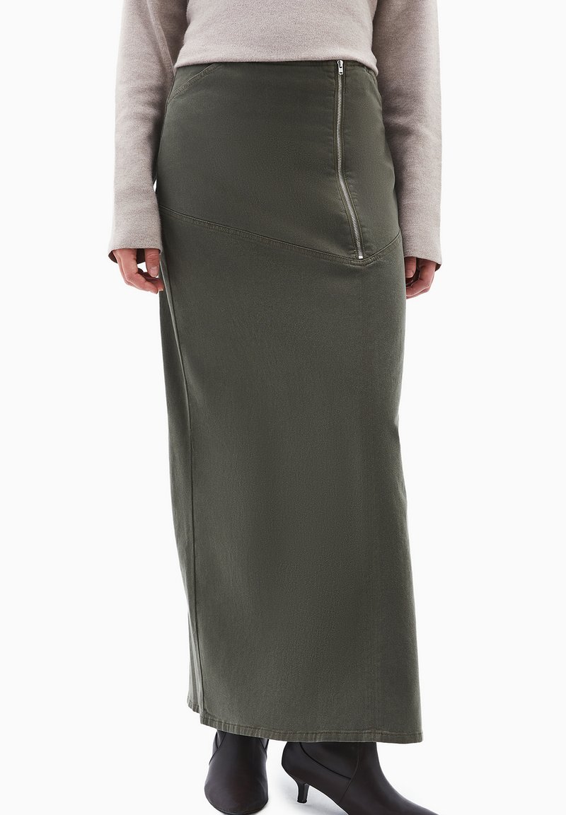 Oliven grøn lang nederdel med en diagonal lynlåsdetalje foran, lavet af tekstureret stof, parret med sorte hælede støvler.