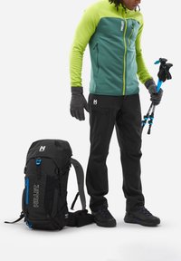 Schwarzer Hirse-Rucksack mit blauen Akzenten, grüne und petrolfarbene Reißverschlussjacke, dunkle Hose, graue Handschuhe und Trekkingstöcke in einer neutralen Umgebung.