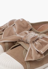 Beige Stoffschuhe mit großen, weichen Samtschleifen oben und weißen, strukturierten Gummikappen an der Spitze, Nahaufnahme.