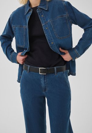 Personne portant une veste en jean bleue et un jean, avec un t-shirt noir et une ceinture noire, les mains posées sur les hanches, vue des épaules aux cuisses.