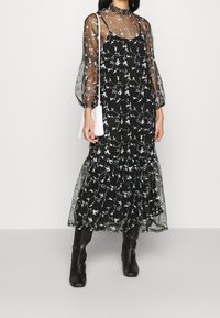 Sort blomster-maksikjole med gennemsigtige, puffede ærmer. Har delikat blå blomstermbroderi og lagdelte flæser ved kanten.