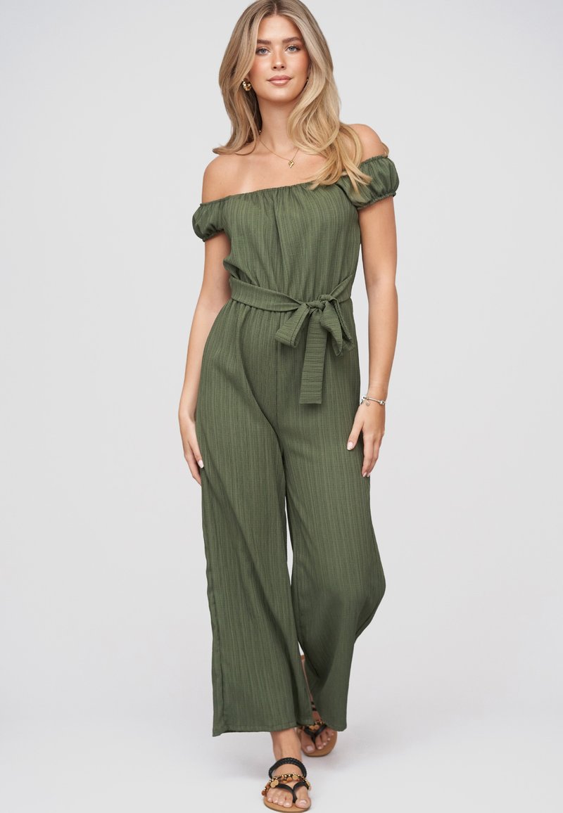Jeune femme aux longs cheveux blonds portant une combinaison vert olive à épaules dénudées avec une ceinture à la taille et des sandales noires, debout devant un fond clair.