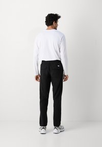 Zwarte broek met achterzakken, elastische tailleband en een wit shirt met lange mouwen. Draagt het met grijs en zwarte sneakers op een effen achtergrond.