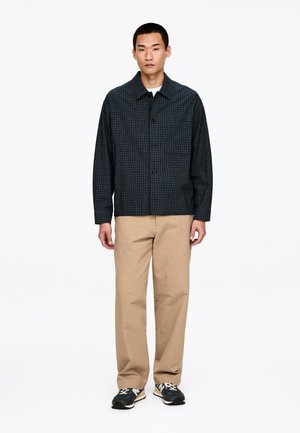Granatowa koszula w kratę z kołnierzykiem, guzikami z przodu i kieszenią na piersi, zestawiona z beżowymi spodniami i czarnymi tenisówkami.