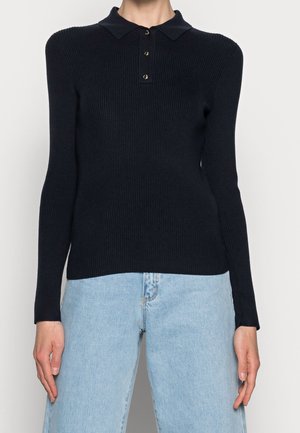 Žena oblečená v přiléhavém černém žebrovaném polotriku s dlouhým rukávem a světle modrých vysokých džínách, stojící pohodlně s rukama u těla.