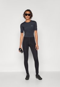 Svart tettsittende topp med korte ermer og høyt liv leggings, laget av glatt, elastisk materiale; modell holder en svart vannflaske, iført solbriller.