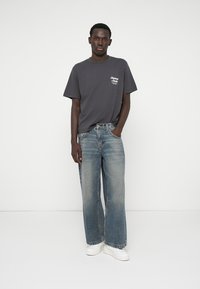 Camiseta cinza de manga curta com estampa de logotipo, combinada com jeans de perna larga em azul claro e ténis brancos. Conjunto simples e casual.