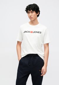 Camiseta blanca de algodón con logo negro "JACK & JONES" en el frente, combinada con pantalones jogger azul marino oscuro que llevan un logo con acento naranja.