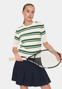 Gestreepte trui met korte mouwen in crème en groen, gecombineerd met een marineblauwe geplooide tennisrok. Houdt een tennisracket vast.