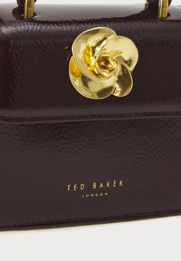 Ted Baker väska i mörk burgunderfärgad lackläder med en texturerad yta, som har ett gyllene rosettförsett lås och präglad logotyp.