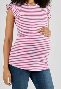 Kobieta w ciąży ubrana w różową bluzkę w białe paski z falbanami i niebieskie dżinsy, z ręką opartą na brzuchu.