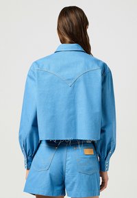 Kort denimjacka i ljusblått, med ett spetsigt okdesign, knäppta manschetter och synliga sömnadsdetaljer. Matchas med matchande shorts.