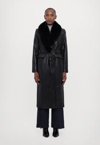 Μαύρο δερμάτινο μακρύ παλτό με ψεύτικη γούνα στον γιακά, λεπτομέρεια με ζώνη και δύο μπροστινές τσέπες. Φοριέται πάνω από φαρδιά μπλε παντελόνια και μπότες μέχρι τον αστράγαλο.