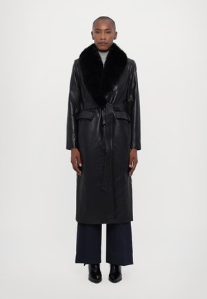 Manteau long en cuir noir avec un col en fausse fourrure, des détails de ceinture et deux poches avant. Porté avec un pantalon large bleu marine et des bottines.