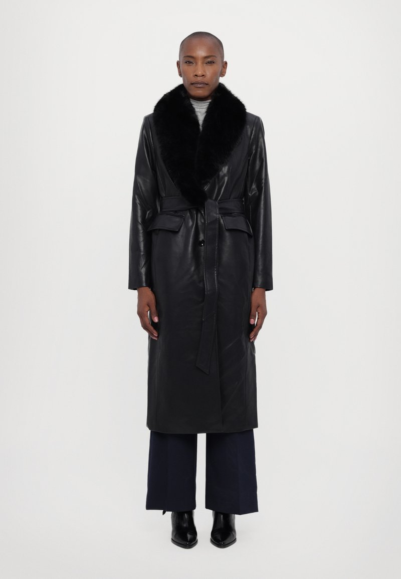 Μαύρο δερμάτινο μακρύ παλτό με ψεύτικη γούνα στον γιακά, λεπτομέρεια με ζώνη και δύο μπροστινές τσέπες. Φοριέται πάνω από φαρδιά μπλε παντελόνια και μπότες μέχρι τον αστράγαλο.