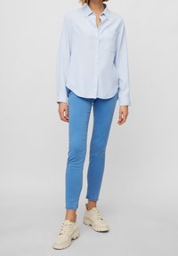 Camicia azzurro chiaro a maniche lunghe con bottoni e taschino sul petto, abbinata a jeans blu attillati e sneakers beige con suola spessa.