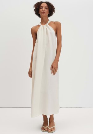 Frau mit lockigem Haar, die ein langes, ärmelloses weißes Neckholderkleid und beige offene Sandalen trägt, stehend vor einem schlichten weißen Hintergrund.