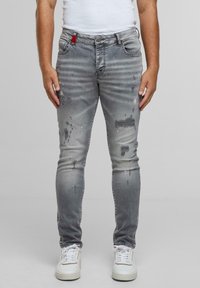 Slim fit szürke farmernadrág, amely kopott részletekkel és kontrasztos piros hurokkal rendelkezik a deréknál. Fehér sportcipővel párosítva.