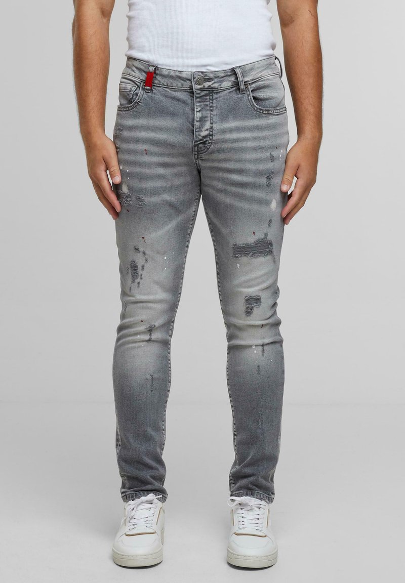 Slim fit szürke farmernadrág, amely kopott részletekkel és kontrasztos piros hurokkal rendelkezik a deréknál. Fehér sportcipővel párosítva.