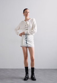 Pull blanc à côtes avec un design lacé, associé à une jupe en denim blanche avec des accents tie-dye bleus, et des bottes lacées noires.