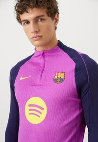 Pink og marineblå atletiktrøje med lynlåshals, der har FC Barcelona-logoet og et stort gult Spotify-logo på fronten.