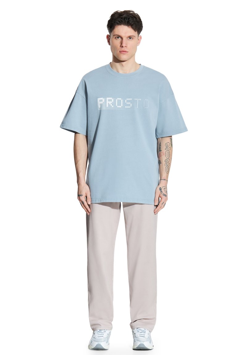 Jasnoniebieska, oversize koszulka z napisem "PROSTO" wyszytym kryształkami, w zestawie z beżowymi, luźnymi spodniami i jasnymi sneakersami.