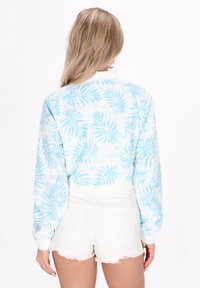 Giacca bianca cropped con motivo di foglie tropicali azzurro chiaro, maniche lunghe e polsini a coste. Abbinata a pantaloni corti bianchi sfilacciati.