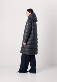 Model staat zijwaarts en toont een lange, donkere pufferjas met capuchon, gecombineerd met wijde jeans, tegen een neutrale achtergrond.