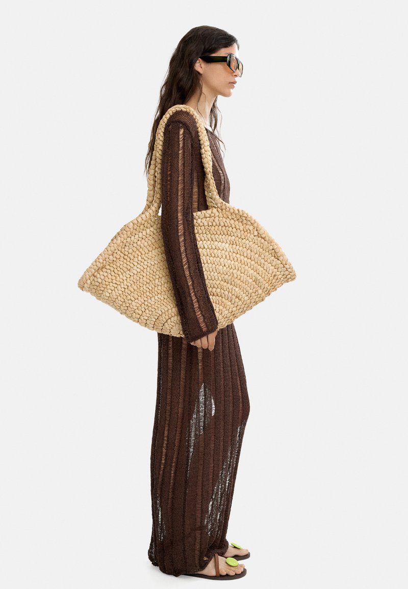 Wpleciona torba ze słomy w naturalnym kolorze o szerokim, trójkątnym kształcie i grubych uchwytach. Nosi się ją z lekką, brązową sukienką dzianinową i sandałami.