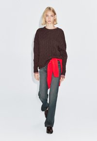 Maglione in maglia marrone, cravatta in tessuto rossa in vita, jeans svasati blu, scarpe marroni, in piedi davanti a uno sfondo grigio chiaro.