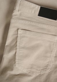 Beige ribfluweelbroek met een achterzak in een rechthoekige vorm en zichtbare stikseldetails. Zwart leren etiket op de tailleband.