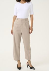 Pantalons beiges ajustés à taille haute, avec un devant plissé et une coupe droite. Associés à un haut blanc ajusté et des chaussures à talons noirs.