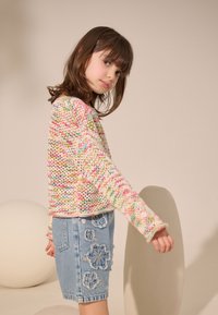 Pull en tricot coloré avec des motifs roses, jaunes et bleus, associé à une jupe en denim déchiré avec des détails de broderie florale.