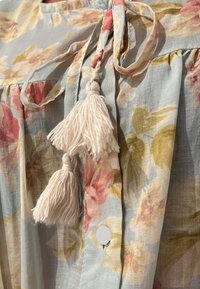 Blusa con motivo floreale in tessuto azzurro chiaro con fiori rosa e gialli. Presenta nastri in tessuto sottili e frange bianche sul collo.