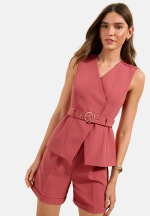 Femme portant un gilet sans manches rose poudré ceinturé et un short assorti à hauteur du genou sur fond blanc.