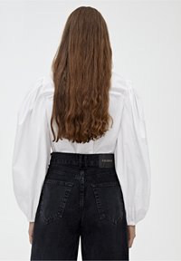 Blusa blanca de mangas abullonadas combinada con jeans negros de cintura alta que presentan un parche de marca y bolsillos traseros. El cabello es ondulado y cae por debajo de los hombros.
