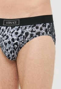 Man wearing black and gray leopard-print briefs with black waistband featuring "VERSACE" logo patch.