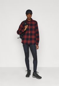 Camisa de flanela em padrão xadrez vermelho e preto, com um bolso no peito, combinada com leggings escuras e botas de caminhada pretas, carregando uma mochila preta.