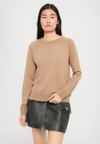 Kvinne som har på seg en beige langermet genser med et lite rosa hjerte og et svart skinn mini nederdel med glidelås foran og lommer.