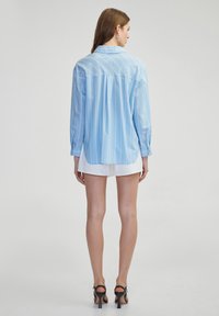 Blusa a rayas azul claro con collar, mangas largas y un dobladillo trasero más largo. Hecha de un tejido suave, presenta rayas verticales blancas.