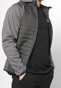 Grå huva jacka med kviltad framdel, släta ärmar och en svart tröja under. Jackan har dragkedja och sidofickor.