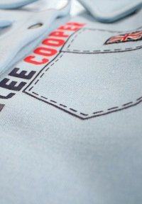 Tissu bleu clair avec lettrage "LEE COOPER" brodé en rouge et noir. Comprend une poche plaquée détaillée avec des surpiqûres contrastées.