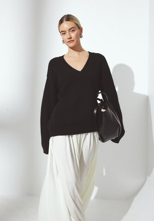 Black wool v-neck sweater with loose fit and wide sleeves, paired with light cream pleated pants and a black leather handbag.
