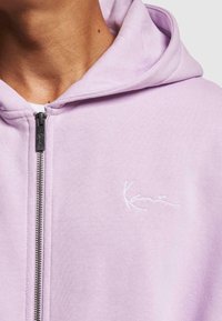 Lilla zip-up hettegenser med kenguru-lomme, ribbede mansjetter og en hvit brodert logo på brystet. Myk bomullsblandingsstoff.