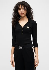 Μαύρο μακρυμάνικο ριπ τοπ με V-λαιμόκοψη, διακοσμημένο με χρυσά κουμπιά και μικρή τσέπη ως ακρόασμα. Φορεμένο με μαύρο παντελόνι με branded ζώνη στη μέση.