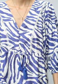 Robe à rayures zèbre bleues et blanches avec un décolleté en V profond, des boutons, des accents en dentelle et une taille à cordon de serrage dotée de franges bleues.