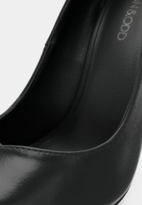 Gros plan sur une chaussure à talon haut en cuir noir montrant une texture lisse et une semelle intérieure incurvée avec une empreinte de marque visible.