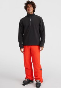 Zwarte fleece pullover met een half-rits kraag, gecombineerd met feloranje skibroek met een losse pasvorm en versterkte stiksels. Zwarte skischoenen.