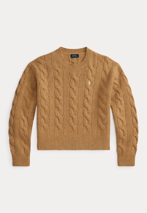 Ung kvinde iført en beige kabelstrikket sweater, løse blå jeans, hvide sneakers og holder en brun ruskindstaske, stående mod en neutral baggrund.